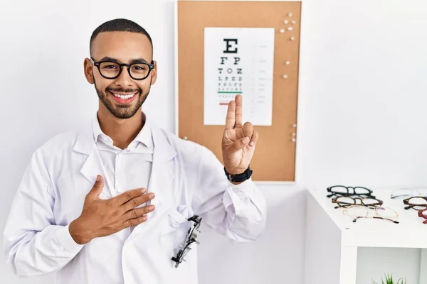 African american optician man standing by eyesight test smiling swearing with hand on chest and fingers up, making a loyalty promise oath
