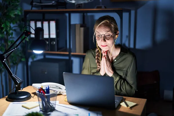 Young blonde woman working at the office at night praying with hands together asking for forgiveness smiling confident.