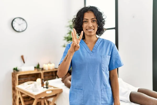 Young therapist woman at wellness spa center showing and pointing up with fingers number three while smiling confident and happy.