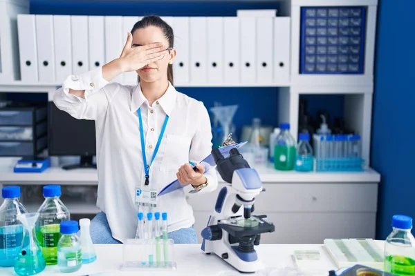 Young brunette woman working at scientist laboratory covering eyes with hand, looking serious and sad. sightless, hiding and rejection concept