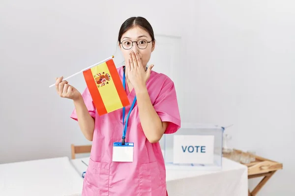 Young asian nurse woman at political campaign election holding spain flag covering mouth with hand, shocked and afraid for mistake. surprised expression