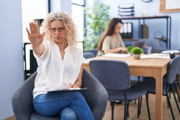 在办公室工作的中年妇女张开手拿着剪贴板 做了个停下来的手势 脸上带着严肃而自信的表情和防御的手势 — 图库照片