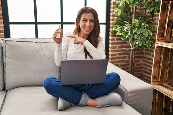 年轻的黑发女人拿着笔记本电脑 手里拿着新家的钥匙 开心地微笑着 手指手画脚 — 图库照片