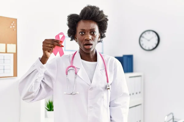 African doctor woman holding pink cancer ribbon at clinic scared and amazed with open mouth for surprise, disbelief face