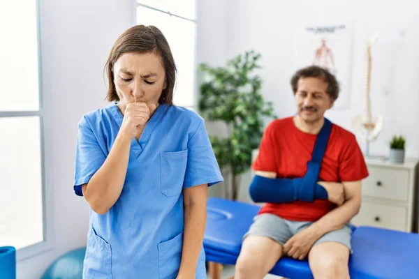 Middle age doctor woman with patient with arm injury at rehabilitation clinic feeling unwell and coughing as symptom for cold or bronchitis. health care concept.