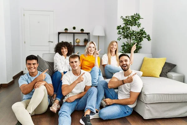 Group of people sitting on the sofa and floor at home smiling and looking at the camera pointing with two hands and fingers to the side.