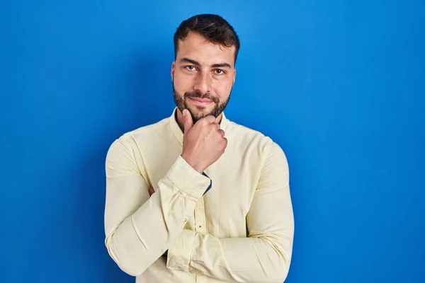 Handsome hispanic man standing over blue background looking confident at the camera smiling with crossed arms and hand raised on chin. thinking positive.