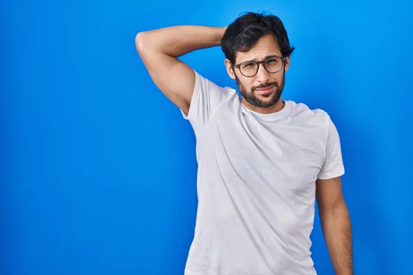 Handsome latin man standing over blue background confuse and wonder about question. uncertain with doubt, thinking with hand on head. pensive concept.