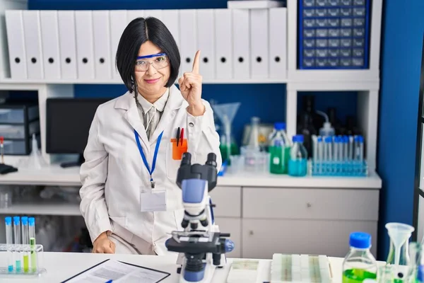 Young asian woman working at scientist laboratory pointing finger up with successful idea. exited and happy. number one.