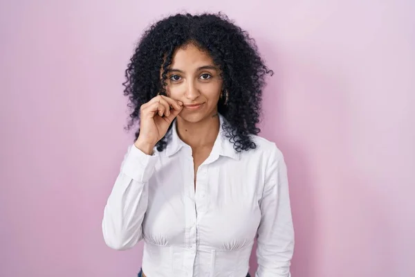 Hispanic woman with curly hair standing over pink background mouth and lips shut as zip with fingers. secret and silent, taboo talking