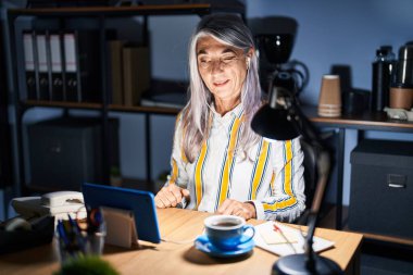 Gri saçlı orta yaşlı bir kadın gece ofiste çalışıyor. Seksi bir ifadeyle kameraya bakıyor. Neşeli ve mutlu bir yüzü var.. 
