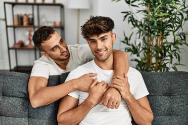 İki İspanyol erkek, kendilerine güvenerek gülümsüyor ve evde birbirlerine sarılıyorlar.