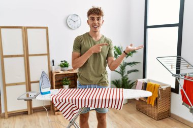 Young caucasian man ironing clothes at home amazed and smiling to the camera while presenting with hand and pointing with finger. 