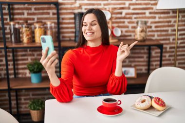 Young hispanic woman eating doughnuts and drinking a cup of coffee doing video call smiling happy pointing with hand and finger to the side 