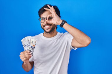 Handsome latin man holding dollars banknotes smiling happy doing ok sign with hand on eye looking through fingers 