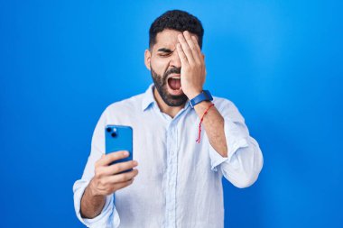 Hispanic man with beard using smartphone typing message yawning tired covering half face, eye and mouth with hand. face hurts in pain. 