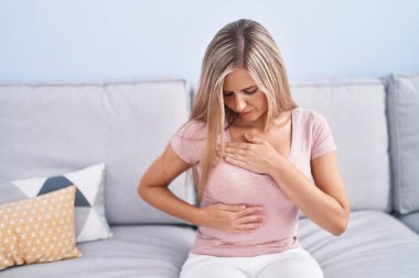 Young blonde woman sitting on sofa examining breast with hands at home