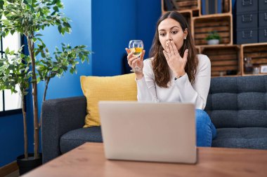 Young hispanic woman doing video call drinking white wine covering mouth with hand, shocked and afraid for mistake. surprised expression 