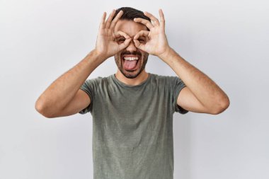 Young hispanic man with beard wearing casual t shirt over white background doing ok gesture like binoculars sticking tongue out, eyes looking through fingers. crazy expression. 