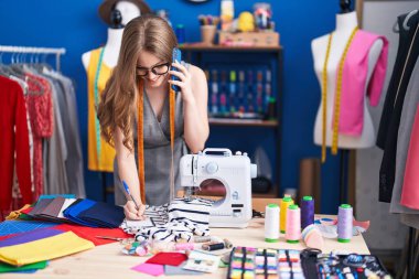 Young woman tailor talking on smartphone writing on notebook at clothing factory