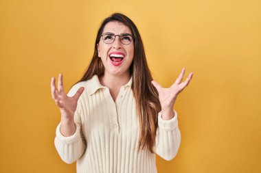 Young hispanic woman standing over yellow background crazy and mad shouting and yelling with aggressive expression and arms raised. frustration concept. 