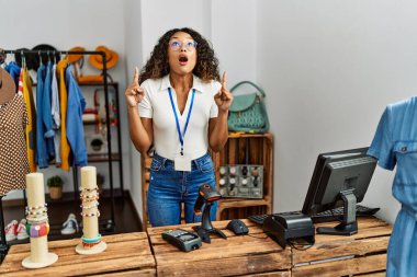 Giyim mağazasında çalışan güzel İspanyol kadın şaşırmış ve şaşırmış bir şekilde yukarı bakıyor ve parmaklarıyla işaret ediyor ve kolunu kaldırıyor.. 