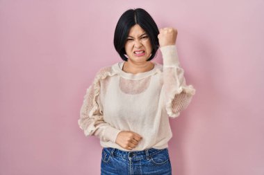 Young asian woman standing over pink background angry and mad raising fist frustrated and furious while shouting with anger. rage and aggressive concept. 