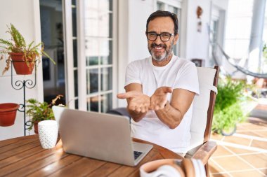 Orta yaşlı bir adam evde dizüstü bilgisayar kullanıyor avuçlarını birbirine değdirip gülümsüyor ve el hareketi yapıyor. Bekleme ve koruma 