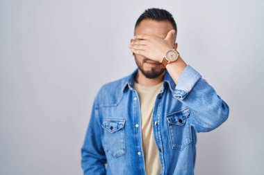 Young hispanic man standing over isolated background covering eyes with hand, looking serious and sad. sightless, hiding and rejection concept 