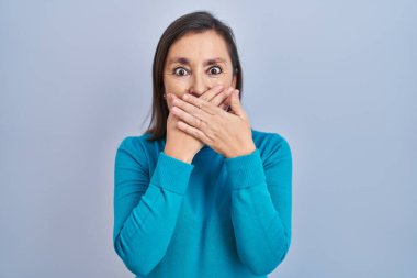 Orta yaşlı İspanyol bir kadın, izole edilmiş arka planda dikiliyor. Ağzını yanlışlıkla ellerinle kapatarak şoka uğruyor. gizli kavram. 