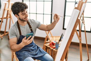 Genç İspanyol ressam mutlu bir resim çiziyor ve resim stüdyosunda akıllı telefon kullanıyor..