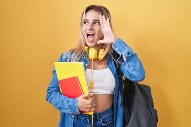Young blonde woman wearing student backpack and holding books shouting and screaming loud to side with hand on mouth. communication concept. 