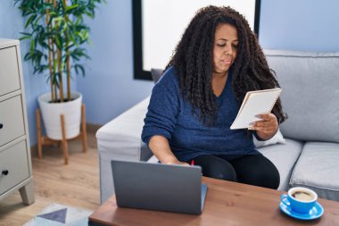 African american woman using laptop reading notebook at home