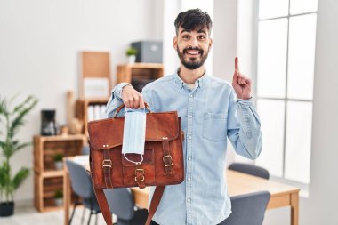 Ofiste sakallı, elinde bavul ve güvenlik maskesi olan genç bir İspanyol adam bir fikir veya soru işareti ile gülümsüyor. 