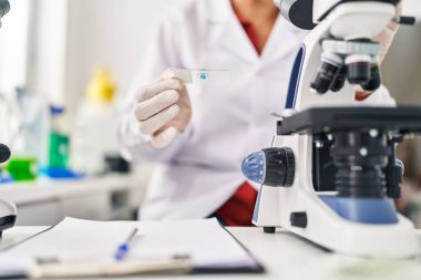 Laboratuvarda mikroskop kullanan orta yaşlı İspanyol kadın bilim adamı üniforması giyiyor.