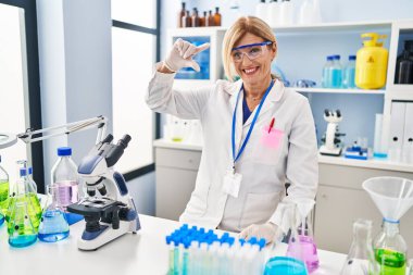 Middle age blonde woman working at scientist laboratory smiling and confident gesturing with hand doing small size sign with fingers looking and the camera. measure concept. 
