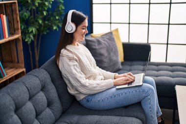 Evdeki kanepede oturan dizüstü bilgisayar ve kulaklık kullanan genç bir kadın.