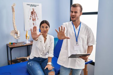 Young hispanic woman at physiotherapist appointment doing stop gesture with hands palms, angry and frustration expression 