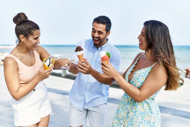 Üç genç İspanyol arkadaş gülümseyerek kumsalda dondurma yiyor..