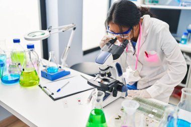 Young chinese woman wearing scientist uniform using microscope at laboratory