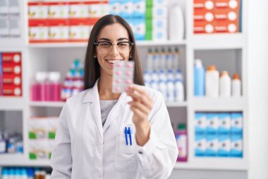 Eczanede kendine güvenen güzel İspanyol kadın eczacı elinde haplarla gülümsüyor.