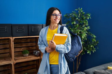 Young hispanic woman student holding book standing at home