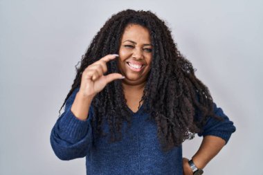 Plus size hispanic woman standing over white background smiling and confident gesturing with hand doing small size sign with fingers looking and the camera. measure concept. 