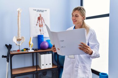 Fizyoterapi kliniğinde fizyoterpist üniforması giyen genç sarışın kadın tıbbi rapor okuyor.