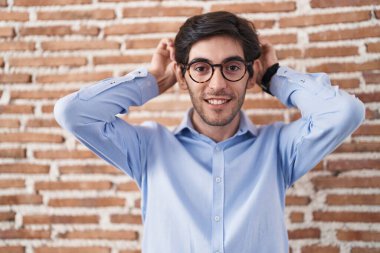 Young hispanic man standing over brick wall background posing funny and crazy with fingers on head as bunny ears, smiling cheerful 