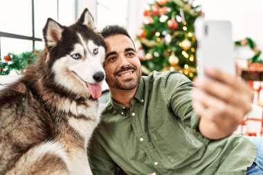 Genç İspanyol adam akıllı telefonuyla selfie çekiyor evdeki yılbaşı ağacında köpekle birlikte kanepede oturuyor.