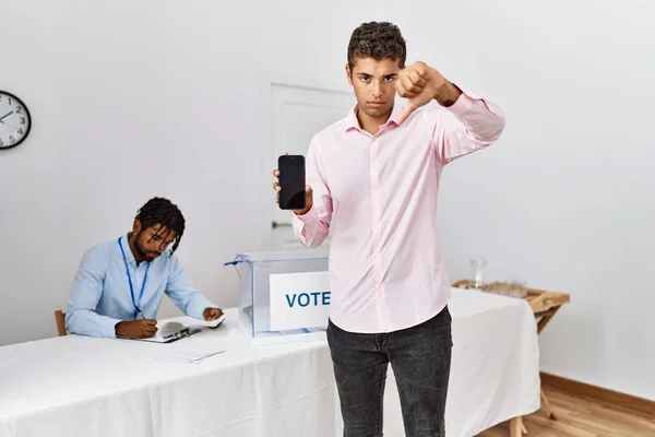 Young hispanic men at political campaign election holding smartphone with angry face, negative sign showing dislike with thumbs down, rejection concept