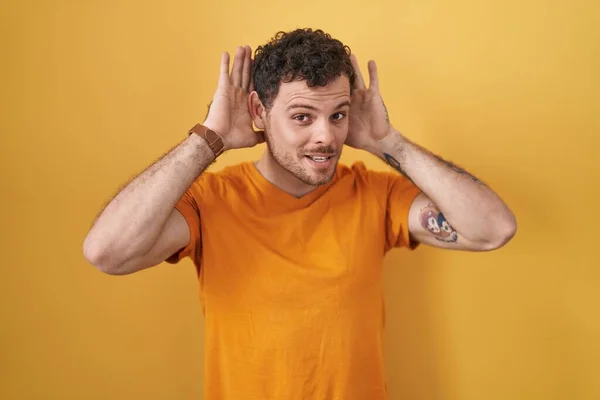 Joven Hombre Hispano Pie Sobre Fondo Amarillo Tratando Escuchar Ambas —  Fotos de Stock