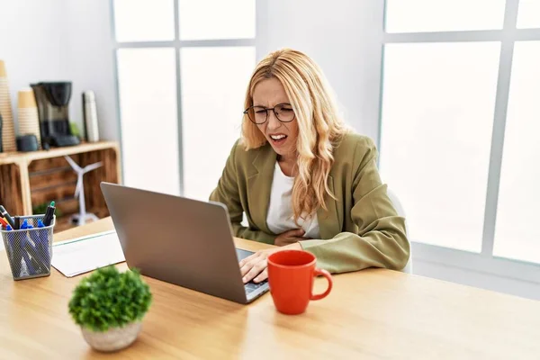 Beautiful blonde woman working at the office with laptop with hand on stomach because indigestion, painful illness feeling unwell. ache concept.