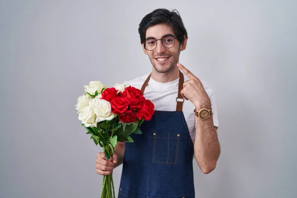 Young hispanic man holding bouquet of white and red roses smiling cheerful showing and pointing with fingers teeth and mouth. dental health concept.